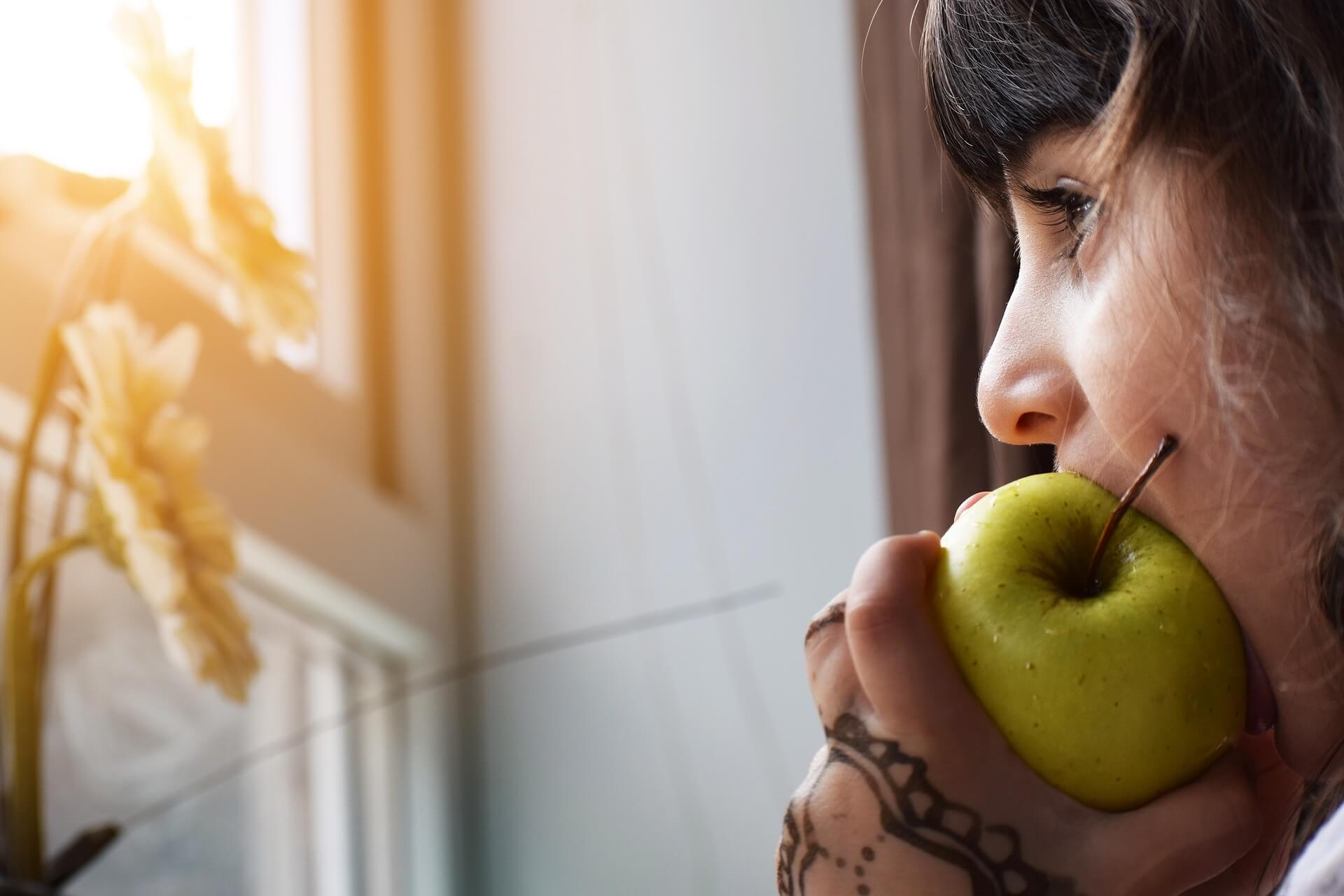 A girl looking at flowers in front of a window and biting a green apple.